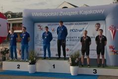  Kajakarze - w trzech dwuosobowych składach - stoją wyprostowani na podium po odebraniu medali. W tle dmuchana ścianka z logotypami partnerów imprezy.